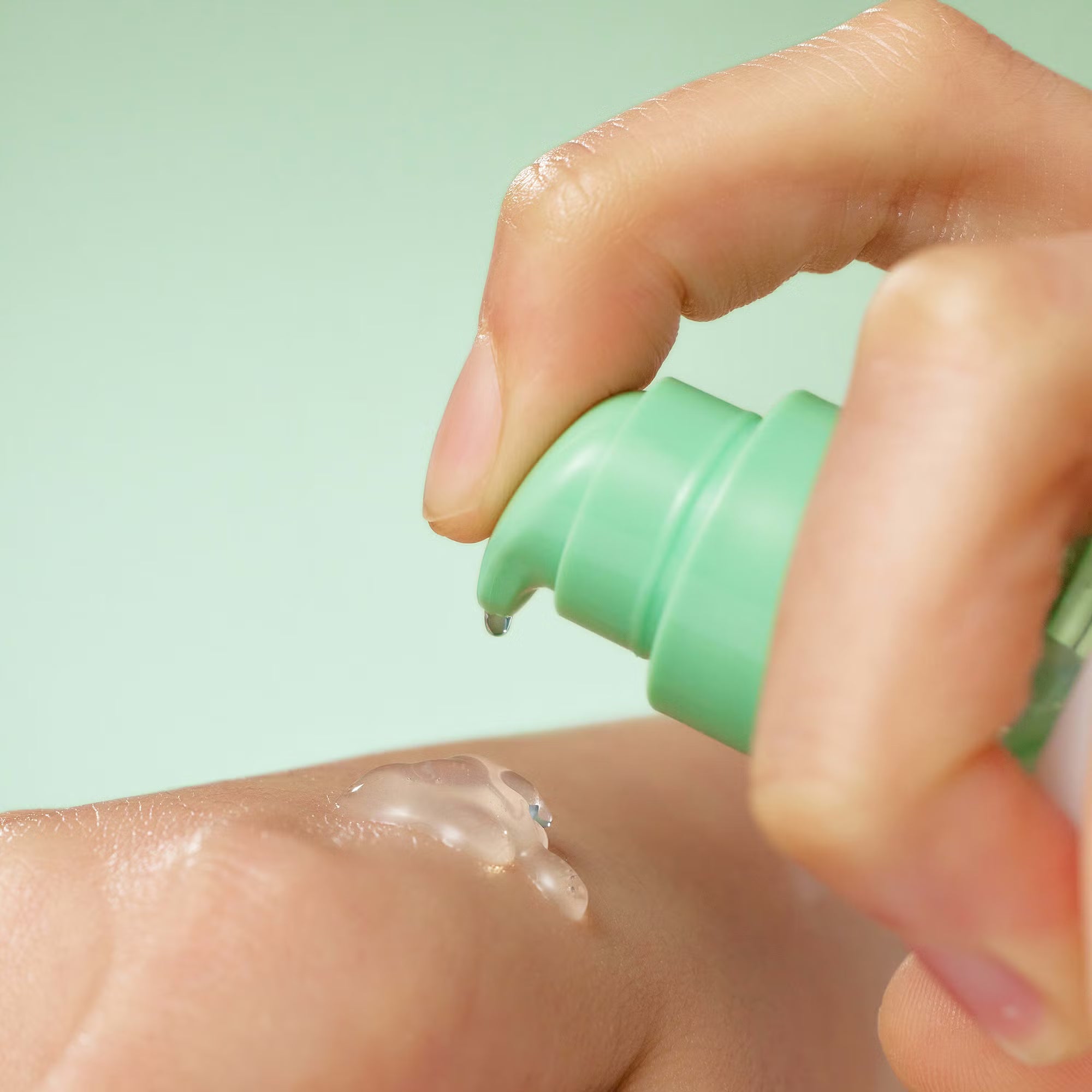 Hand applying a clear cream from a green bottle onto skin against a light green background