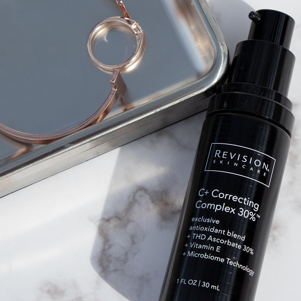 Revision Skincare C+ Correcting Complex 30% bottle on a marble surface with a mirror and ring in the background.